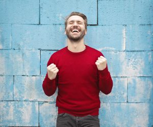 Mężczyzna stoi przed ścianą. ma zamknięte oczy, uniesione ręce i zaciśnięte pięści. Jest zadowolony i bardzo się cieszy.