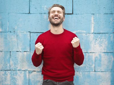 Mężczyzna stoi przed ścianą. ma zamknięte oczy, uniesione ręce i zaciśnięte pięści. Jest zadowolony i bardzo się cieszy.