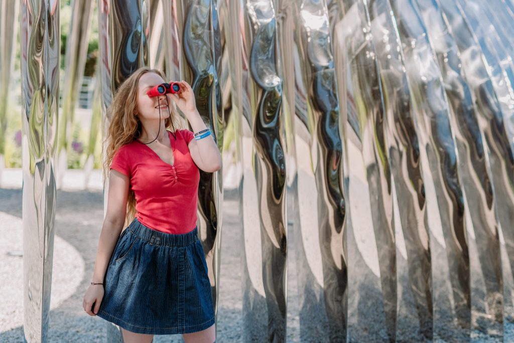 Młoda dziewczyna stoi przed szklaną ścianą. Patrzy w dal przez małą lornetkę przyłożoną do oczu. Jest uśmiechnięta.