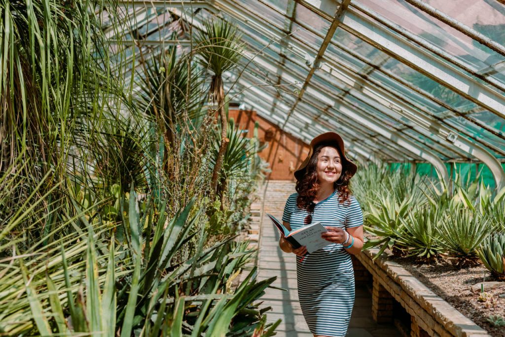 Młoda uśmiechnięta dziewczyna w sukience i kapeluszu na głowie. Patrzy w dal, trzyma książkę w ręce. Jest w oszklonej oranżerii pełnej roślin.