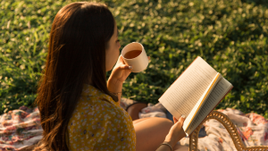 Młoda dziewczyna siedząca na trawie i czytająca książkę. W lewej ręce trzyma kubek z kawą.