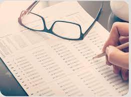 Biała kartka papieru na stole z pytaniami testowymi. Na kartce położone okulary. Widać fragment dłoni trzymającej ołówek. Dłoń pochylona nad kartką.