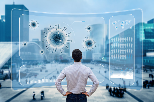 Mężczyzna stojący tyłem z podpartymi rękami. Patrzy na duży, przeszklony ekran z wykresami. W tle szyba i widoczne przez nią biurowce i ludzie na ulicy.