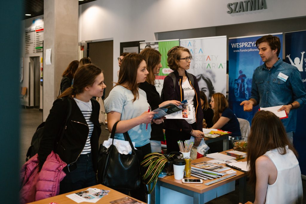 Trzy studentki stoją przy stoisku pracodawcy poczczas targów pracy.