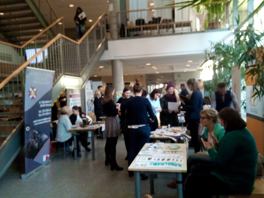 Duży korytarz i schody jednego z budynków uczelni, a w nim grupa młodych ludzi stojąca przy różnych stoiskach pracodawców podczas targów pracy.