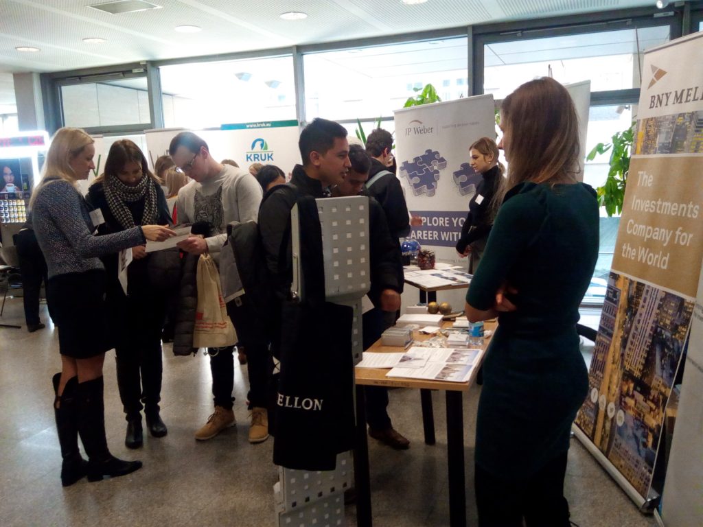 Duży korytarz a w nim grupa młodych ludzi stojąca przy różnych stoiskach pracodawców podczas targów pracy.