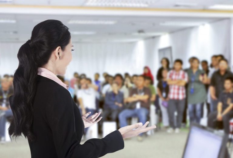 Młoda kobieta w czarnej marynarce prowadzi prezentację w sali. W tle widać grupę osób patrzących w stronę kobiety.