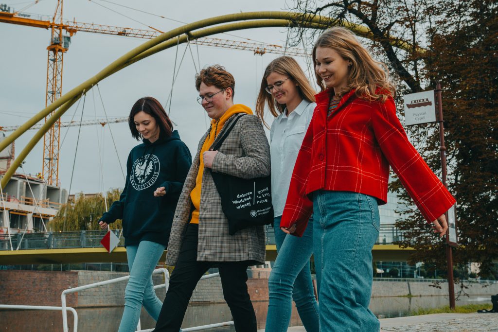 Czwórka młodych osób; trzy dziewczyny i chłopak schodzą po schodach. W tle most.