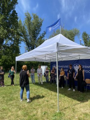 Grupa osób podczas warsztatów stoi pod namiotem z logiem Uniwersytetu Wrocławskiego