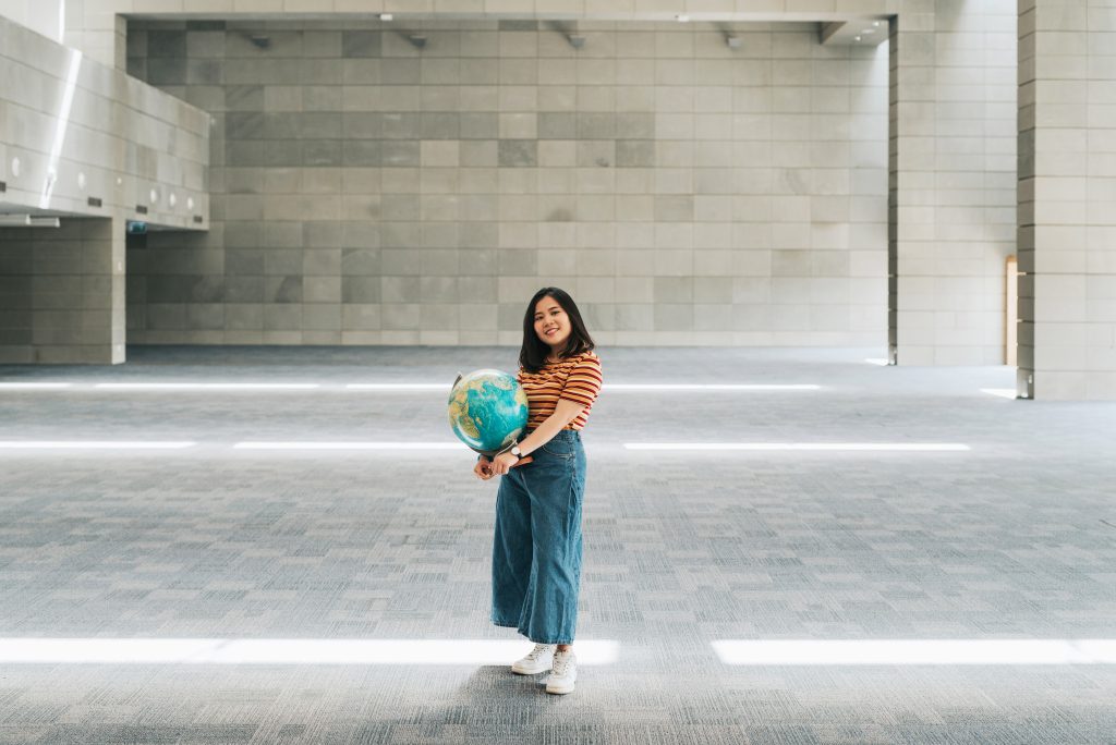 Studentka z globusem w dłoniach stoi pośrodku dużego, jasnego korytarza w bibliotece