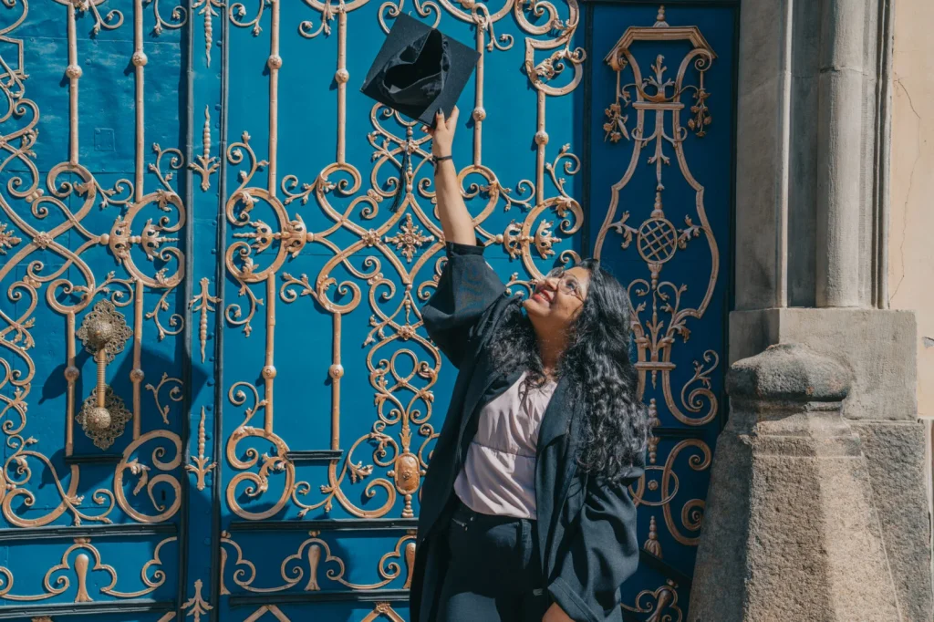 Absolwentka na tle drzwi prowadzących do Gmachu Głównego Uniwersytetu Wrocławskiego. Unosi w ręce biret i się uśmiecha.