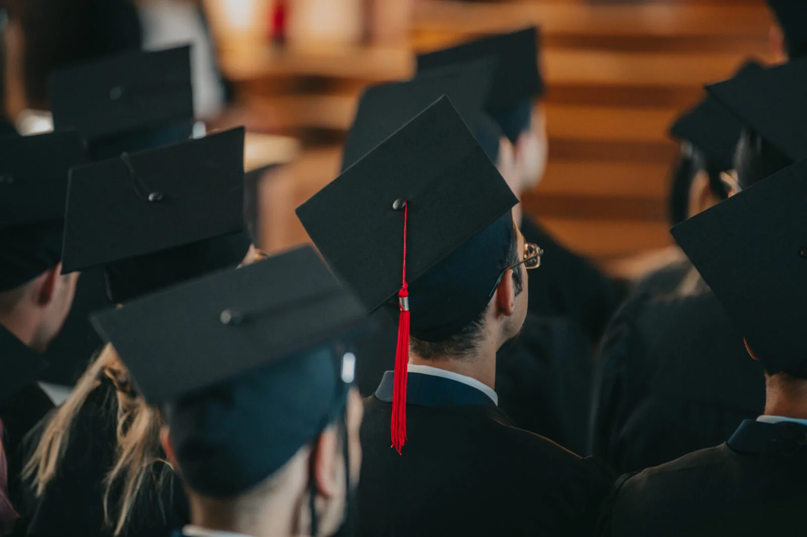 Studenci podczas zakończenia studiów siedzą tyłem w biretach absolwenckich na głowach.