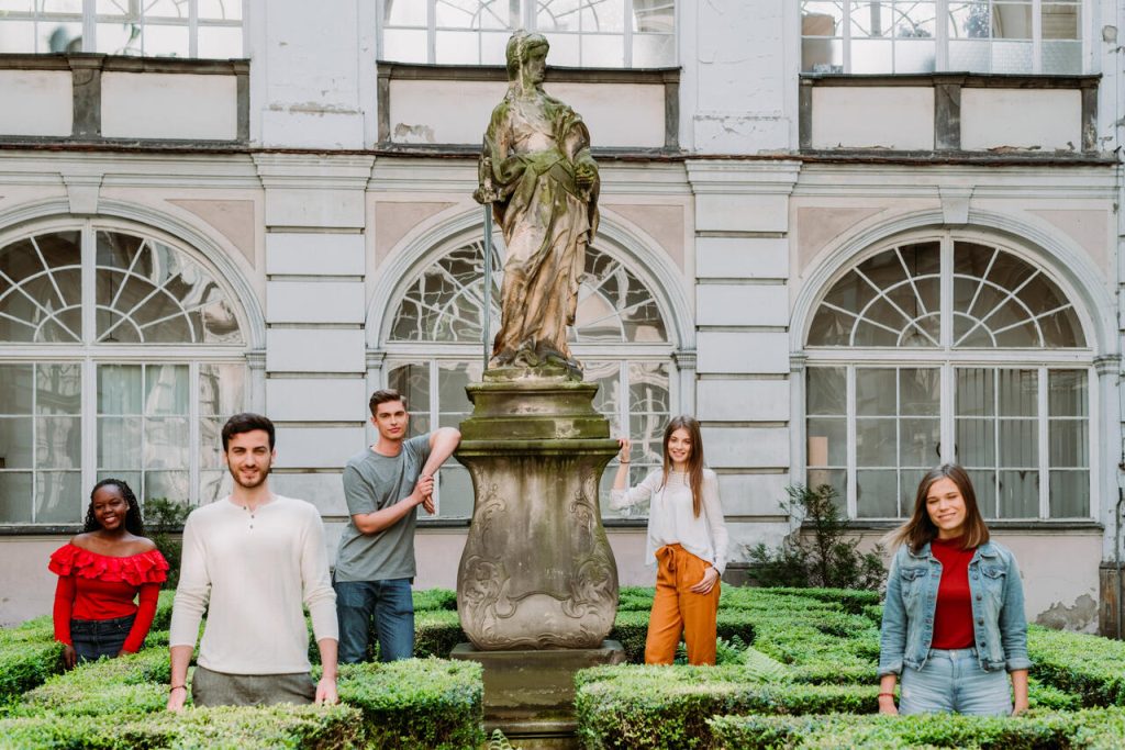 Młodzi ludzie stojący wokół omszałego pomnika kobiety pomiędzy krzewami bukszpanu