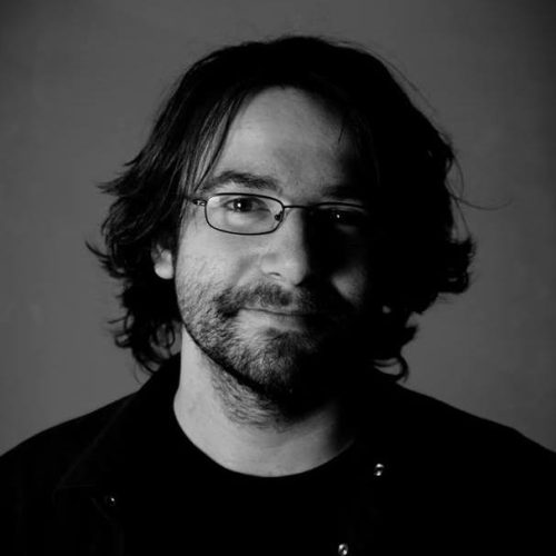 Czarno-białe zdjęcie, portret mężczyzny w dłuższych, ciemnych włosach z okularami.